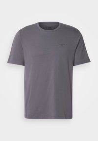 Camiseta de algodón gris con mangas cortas, escote redondo y un pequeño logo negro en el pecho izquierdo. Textura suave, diseño minimalista.