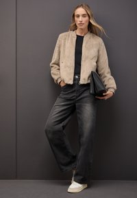 Faux fur bomberjacka i beige, svart topp, vida slitna svarta jeans och vita sneakers med grå detaljer, håller en svart kuvertväska.