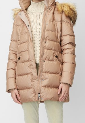 Femme portant un manteau d’hiver beige matelassé avec capuche bordée de fourrure, un pull en laine crème et un pantalon de couleur claire.