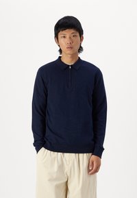 Polo shirt blu navy con zip e colletto, realizzato in un tessuto morbido. Presenta polsini e orlo a costine, abbinato a pantaloni beige in veste rilassata.