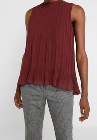 Burgundyröd ärmlös blus med vertikala veck, rund halsringning och en utsvängd nederkant, parat med grå herringbone-byxor.