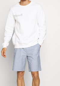 Mand iført hvid langærmet sweatshirt med lille tekst på brystet og lyseblå stribede shorts, stående med den ene hånd i lommen.