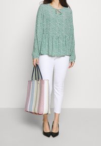 Blusa verde con stampa floreale, collo rotondo e dettaglio con fiocco, abbinata a pantaloni bianchi aderenti e una borsa a tracolla a righe multicolori.