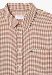 Camisa de cuadros marrón con cuello abotonado y botones blancos. Presenta un bolsillo en el pecho con el logo verde de Lacoste. Diseño de corte regular.