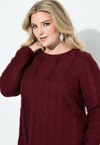 Donna bionda con trucco leggero che indossa orecchini d'argento e un maglione a girocollo color borgogna, in posa con una mano vicino al collo.