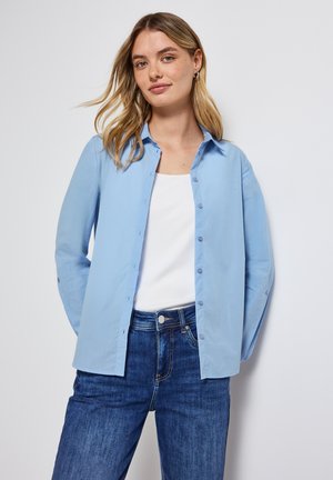 Helles blaues Hemd mit knöpfbarem Kragen aus glattem Stoff, lange Ärmel und kombiniert mit dunkelblauen Jeans.
