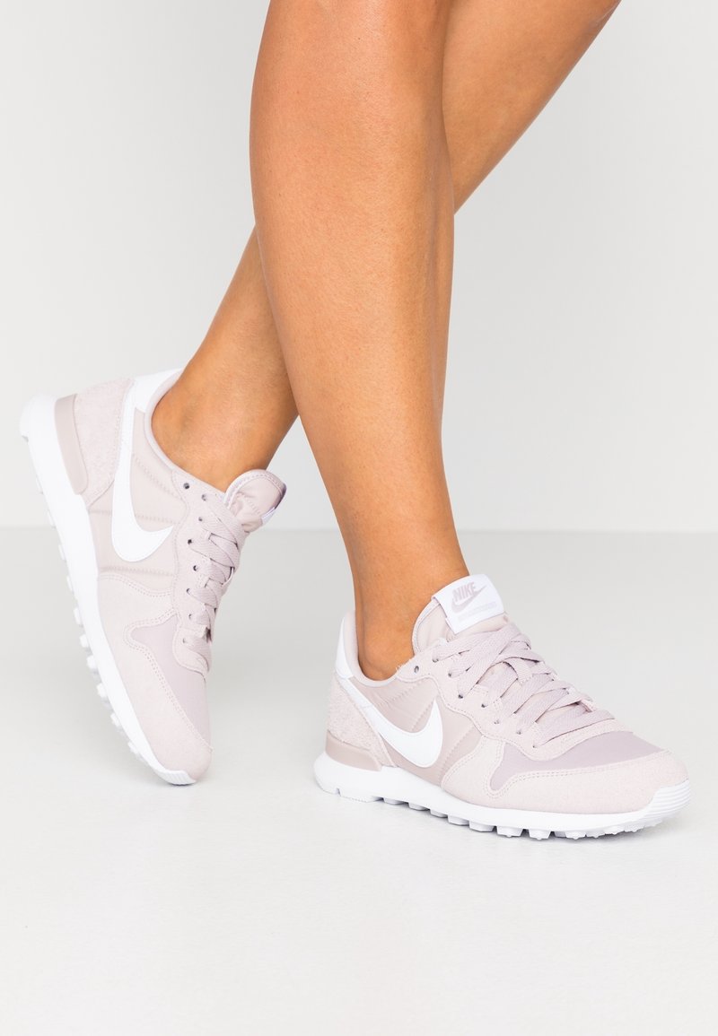 Jambes portant des baskets Nike rose clair avec le logo swoosh blanc, debout sur une surface gris clair unie.