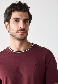 Camiseta de algodón burdeos con cuello redondo, que cuenta con un ribete de rayas en blanco y negro y un pequeño logo bordado en el pecho.