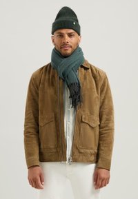 Muž v hnědé semišové bundě, modrém šále, tmavě zelené pletené čepici, světlé košili a bílých kalhotách, stojící na pozadí světle jednolité barvy.