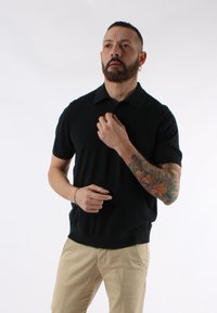 Maglietta polo nera a maniche corte con colletto, orlo arrotondato e una texture morbida. Pantaloni beige. Visibile un notevole tatuaggio colorato su un braccio.