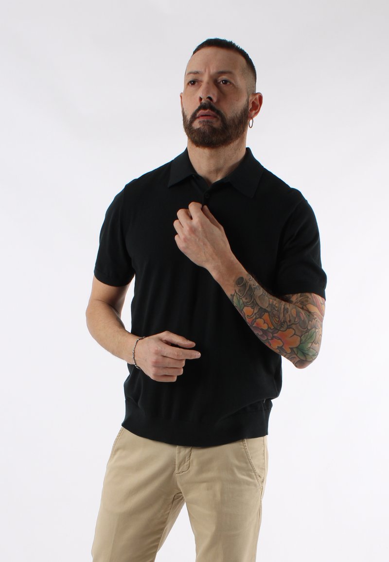 Maglietta polo nera a maniche corte con colletto, orlo arrotondato e una texture morbida. Pantaloni beige. Visibile un notevole tatuaggio colorato su un braccio.