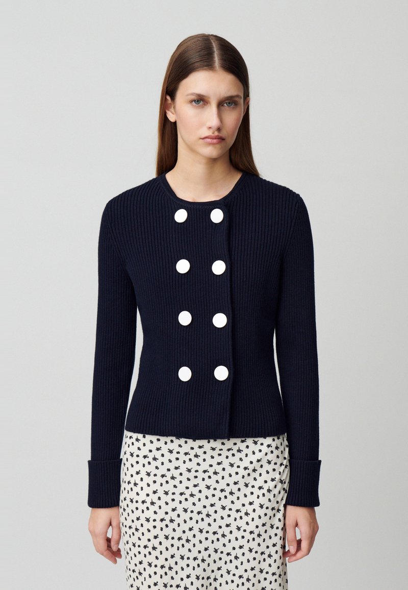 Gilet côtelé marine avec un col rond, des accents de boutons blancs et des manches longues. Associé à une jupe à motifs présentant de petits designs floraux.