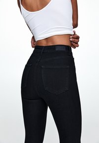 Jean noir à coupe ajustée, avec deux poches arrière et une étiquette de marque sur la ceinture, en denim à texture lisse.