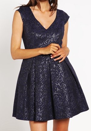 Femme portant une robe bleu marine sans manches avec un décolleté en V et un motif texturé métallique, debout les mains doucement jointes à la taille.