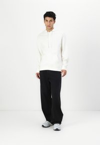Hanorac alb cu buzunar frontal și șnururi, purtat cu pantaloni largi negri și adidași gri. Design și culori simple, casual.