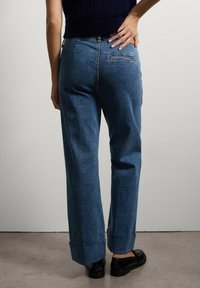Pantalones de mezclilla de pierna ancha en un lavado azul claro. Presentan un dobladillo enrollado, un bolsillo trasero y una textura suave. Combinados con zapatos negros brillantes.