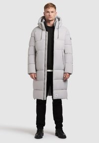 Hellgraue Daunenjacke mit Kapuze, ausgestattet mit einem Reißverschluss vorne, großen rechteckigen Paneelen und Seitentaschen. Getragen über einem schwarzen Oberteil und einer Hose.