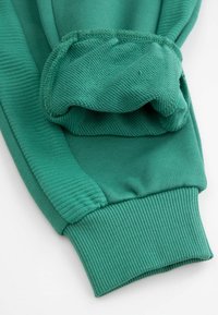 Rękaw zielonej bluzy z prążkowanymi mankietami i teksturowaną powierzchnią. Tkanina wydaje się gładka z widocznymi detalami przeszyć.