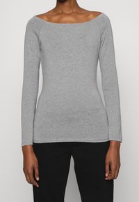 Kvinna som bär en figursydd ljusgrå långärmad tröja med bred båthalsning och svarta byxor, stående mot en enfärgad bakgrund.