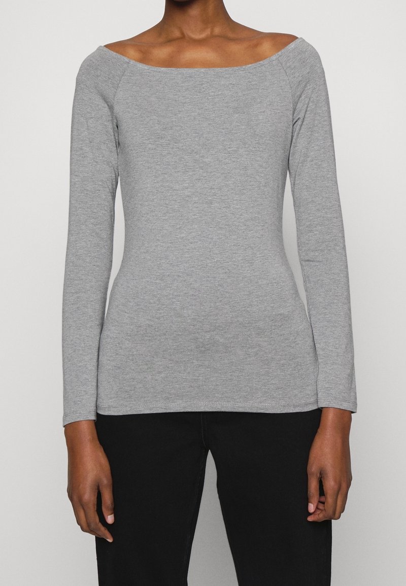 Kvinna som bär en figursydd ljusgrå långärmad tröja med bred båthalsning och svarta byxor, stående mot en enfärgad bakgrund.