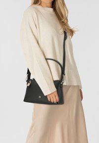 Borsa a mano in pelle nera con finitura testurizzata, dotata di una tracolla lunga, un manico superiore e un dettaglio con lucchetto argentato. Indossata sopra un maglione chiaro.