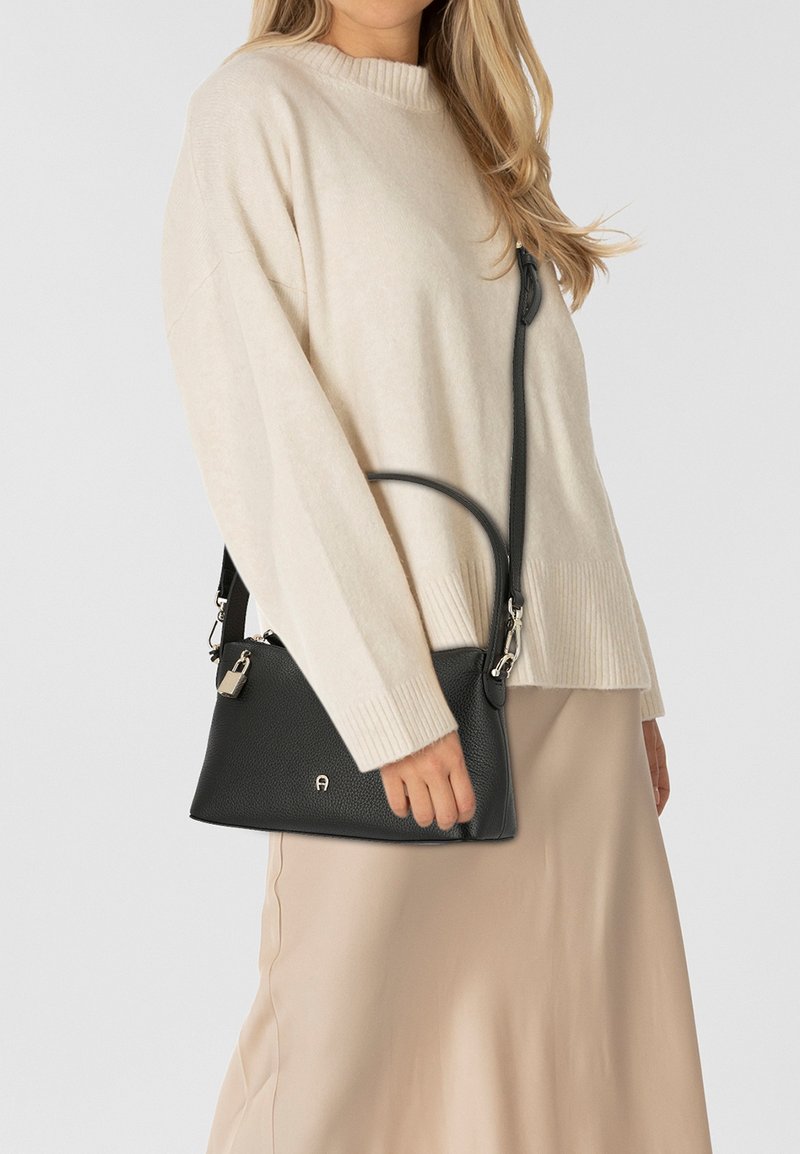 Borsa a mano in pelle nera con finitura testurizzata, dotata di una tracolla lunga, un manico superiore e un dettaglio con lucchetto argentato. Indossata sopra un maglione chiaro.
