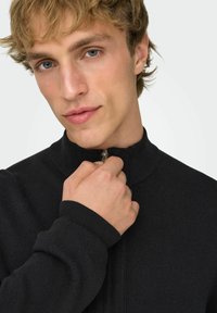 Maglione nero con zip, colletto alto e polsini a costine. Il tessuto sembra strutturato, con una finitura liscia e un design minimalista.