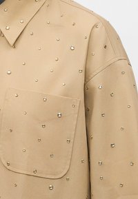 Camisa beige con bolsillo frontal, decorada con pequeñas tachuelas redondas de oro y plata distribuidas uniformemente por toda la tela y el cuello.