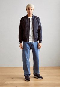 Giubbotto bomber blu navy con polsini a coste, tasca con zip sulla manica, t-shirt bianca, jeans dritti azzurri chiari e sneaker blu navy.