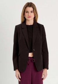 Blazer noir ajusté avec une fermeture à un bouton, des revers à cran et deux poches. Porté sur un top court noir et un pantalon bordeaux.