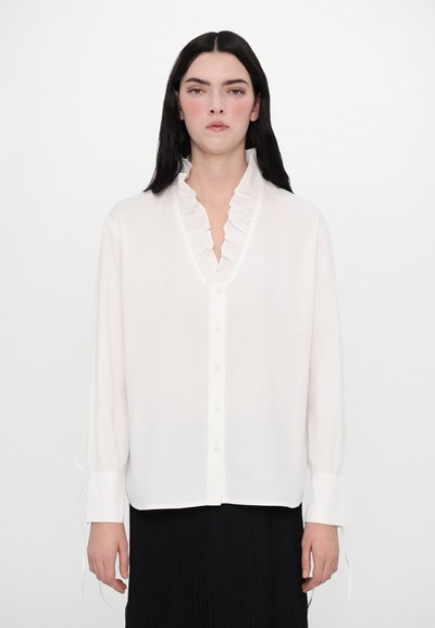 Blouse blanche à manches longues avec un col en V froncé, fermeture à boutons et poignets élastiques. Fabriquée en tissu léger avec une texture lisse.