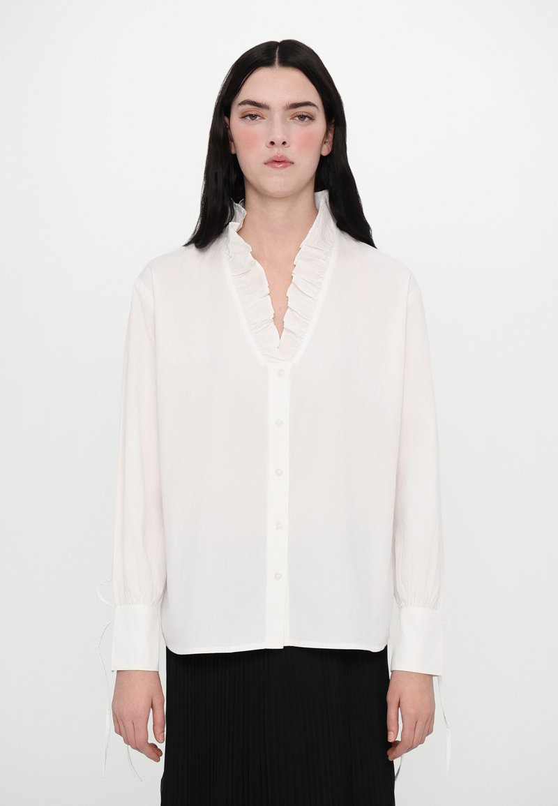 Weiße Bluse mit langen Ärmeln, Rüschenausschnitt, Knopfleiste und elastischen Bündchen. Aus leichtem Stoff mit glatter Textur hergestellt.