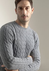 Suéter gris de punto trenzado con cuello redondo y patrón texturizado. Presenta mangas largas, puños acanalados y una silueta ajustada.