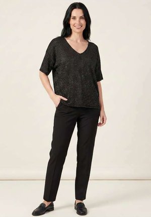 Femme debout, portant un haut noir à col en V et manches courtes, pantalon droit noir et mocassins noirs, souriante avec une main dans la poche.
