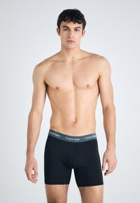 Fekete boxer alsónadrág szürke gumival, amelyen a "Calvin Klein" logó található. Sima anyag, testhezálló kialakítás, combközép hosszal.