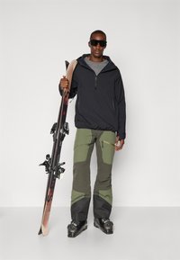Costum de schi compus dintr-un pulover negru cu glugă, un strat de bază gri, pantaloni de schi verzi și gri, și cizme de schi negre, ținând schiuri cu legături.
