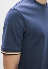 Tengerészkék rövid ujjú póló puha pamutból, bordázott mandzsettákkal, világosbarna, fehér és fekete csíkokkal. Simas textúra és testhezálló szabás.