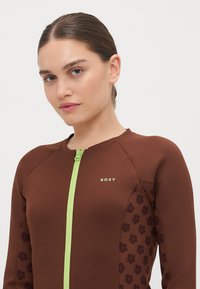 Ruda wetsuit su žaliu užtrauktuku, dekoruotas gėlių raštais šonuose. Pagamintas iš lygios, tamprios medžiagos. Rodo "Roxy" logotipą auksinės spalvos.
