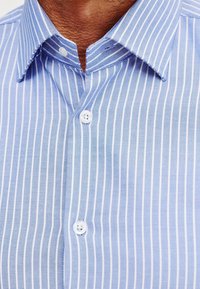 Jasnoniebieska koszula zapinana na guziki z wąskimi białymi paskami, posiadająca klasyczny kołnierzyk i dwa białe guziki z przodu. Miękka tekstura materiału.