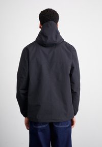 Hombre con una chaqueta con capucha negra y jeans azul oscuro, de pie de espaldas contra un fondo blanco liso.