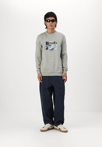 Lys grå hettegenser med grafisk trykk i midten, kombinert med løstsittende marineblå bukser og hvite joggesko med brune detaljer.