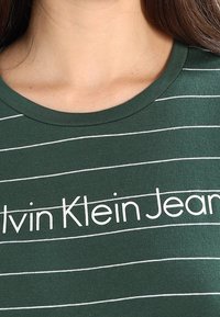 Grünes Baumwoll-T-Shirt mit horizontalen weißen Streifen und einem großen "Calvin Klein Jeans"-Logo in der Mitte. Rundhalsausschnitt und sanfte Textur.