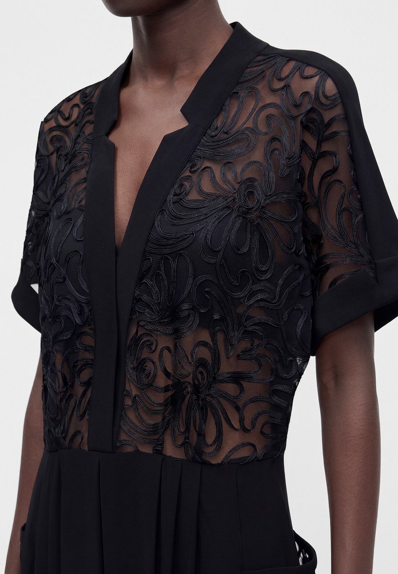 Blouse noire transparente avec broderie florale, manches courtes, col en V profond avec col et poignets noirs unis portée par une personne à la peau foncée.