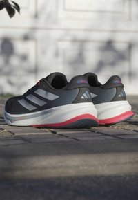 Zapatillas deportivas negras con detalles en gris, parte superior texturizada, una media suela blanca y una suela roja. Presentan tres rayas blancas y un detalle del logo.