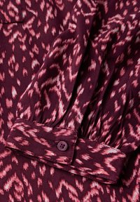 Blouse en tissu bourgogne avec un motif abstrait rose, détails de manches froncées et un bouton violet sur le poignet. Texture douce.