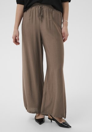 Žena nosi voľné hnedé nohavice so šnúrkou v páse a čierne nízke lodičky so špicatou špičkou na jednoduchom pozadí.