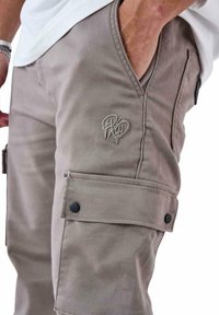 Pantalones cargo beige fabricados con tejido duradero, con un bolsillo con cremallera y un detalle de bordado en el muslo. Incluye acentos de botón negro.