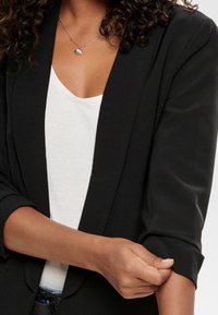Blazer negro con mangas dobladas, combinado con una blusa blanca. El material parece suave con un diseño estructurado y entallado. Detalles mínimos.
