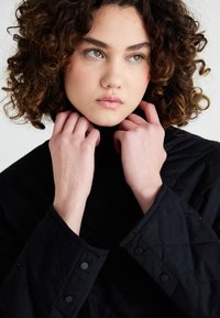Jachetă neagră matlasată cu guler înalt și închideri cu butoane prin presiune, purtată de un model cu păr creț, cu mâinile aproape de gât.