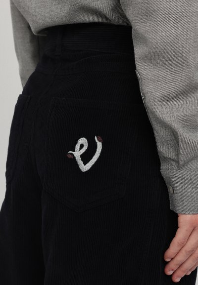 Pantaloni negri din catifea cu un buzunar la spate cu litere brodate albe și accente roșii mici. Partea de sus este gri, având un aspect texturat.